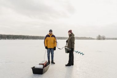 Two adults standing on a frozen lake with ice fishing gear during winter.